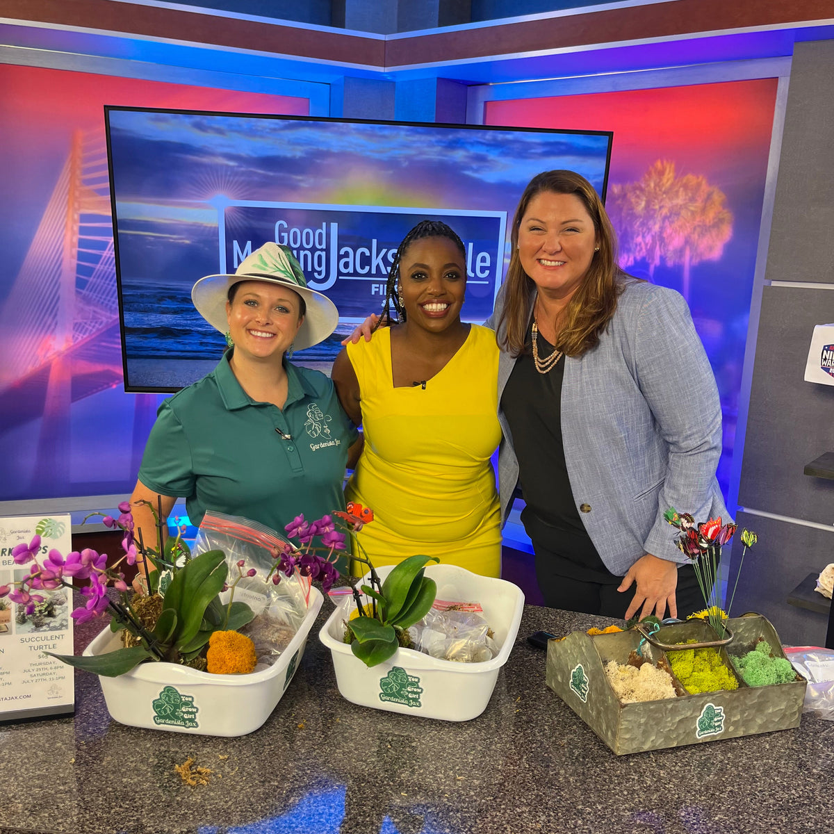 India standing side by side next to two news casters on the good morning Jacksonville show it plants on a table in front of them. 